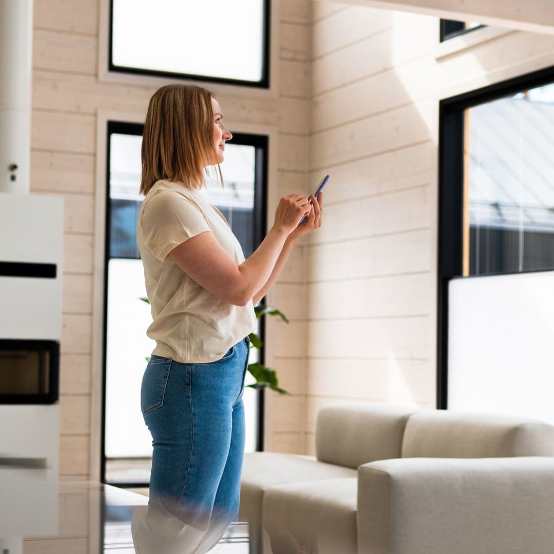 A woman standing in a modern living room uses a smartphone to control motorised window blinds.
